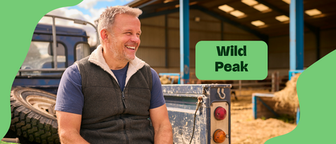 Smiling middle-aged farmer wearing a fleece-lined vest, leaning against the back of a blue truck, with "Wild Peak" text overlayed on a green shape, with a barn visible in the background.