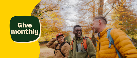Three diverse hikers smiling and walking in a forest with autumn foliage; a green graphic with white text saying "Give monthly" is on the left side.