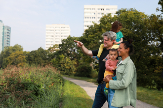 A family of four are at a nature reserve with buildings visible in the background. The father is pointing to show his partner and children something in the distance