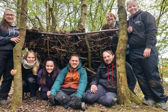 four people sat in a shelter made of sticks with three other stood either side, all are smiling and laughing