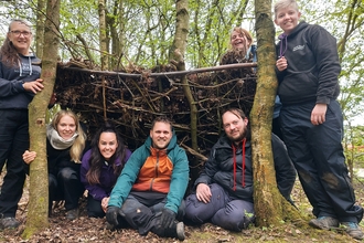 a group of adults stood around and sat under a shelter made of branches and sticks between two trees