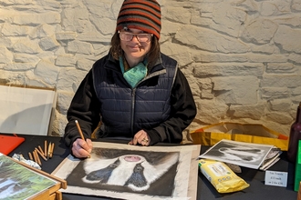 a lady in glasses and a stripy hat creating artwork of a badger smiling at the camera