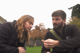 A man and woman crouching on a grassy area in front of a church, the man holding a small orange mushroom and talking to the woman, whilst she inspects the fungi.