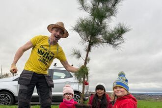 A man and three young girls plant a small tree in a field, under a cloudy sky. The man wears a yellow t-shirt with "One of the Wild Ones" printed on it, work pants, a straw hat, and gloves. The girls wear red hoodies, with hats, and are kneeling by the tree. A car is parked nearby.