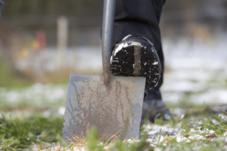 Close-up view of a metal shovel blade on the ground, with a black work boot pressing down on it. The boot tread has visible snow. The shovel blade is slightly rusted, and the ground is covered in patches of snow and green grass. The scene suggests outdoor work in a cold environment.
