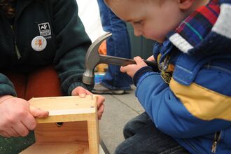 A young boy in a blue jacket carefully uses a hammer to attach a piece of wood to a wooden birdhouse, assisted by an adult holding the birdhouse, likely in an outdoor workshop setting.