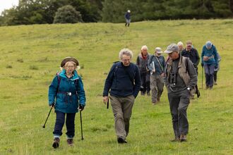 A group of people hike across a grassy field, some with walking sticks and backpacks.