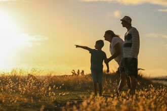 Silhouettes of a family of three in a field of golden grass at sunset, with the child pointing towards the horizon.