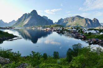 A picturesque coastal village with red and white buildings nestled at the base of dramatic, green-covered mountains, reflected in the calm, blue water.