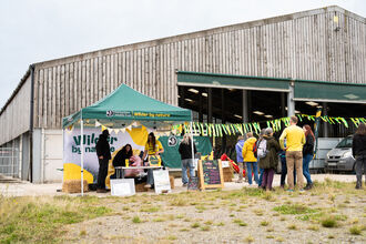 An outdoor event tent for Derbyshire Wildlife Trust, with the slogan "Wilder by nature," is set up in front of a large wooden building, with several people milling around the tent and a car parked in the background.
