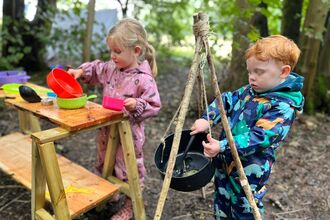 Two young children are engaged in outdoor play, with one pouring water from a red bowl into a green bowl on a wooden table, and the other stirring something in a black pot hanging from a tripod of sticks.