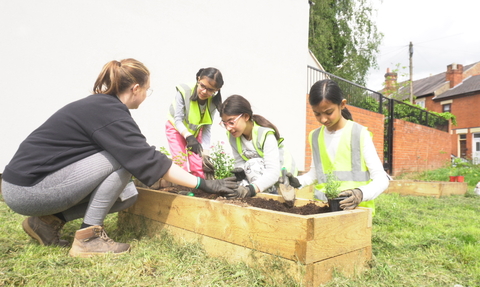 an adult and three children wearing high vis vests are crouched around a raised planter bed planting plants