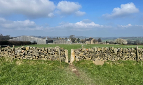 the view of farm buildings in the background with a dry stone wall in the foreground and a blue sky with a couple of clouds