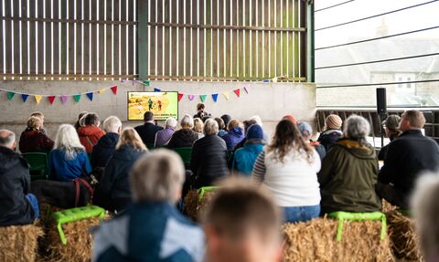 People seated on hay bales listen to a speaker at a meeting inside a barn; a screen displays a logo and image, and colorful pennants hang across the wall.