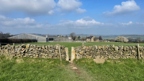 the view of farm buildings in the background with a dry stone wall in the foreground and a blue sky with a couple of clouds