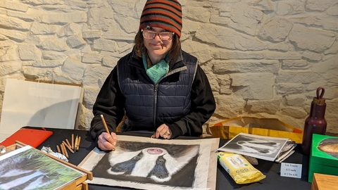a lady in glasses and a stripy hat creating artwork of a badger smiling at the camera