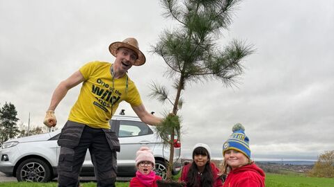 A man and three young girls plant a small tree in a field, under a cloudy sky. The man wears a yellow t-shirt with "One of the Wild Ones" printed on it, work pants, a straw hat, and gloves. The girls wear red hoodies, with hats, and are kneeling by the tree. A car is parked nearby.