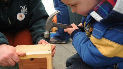 A young boy in a blue jacket carefully uses a hammer to attach a piece of wood to a wooden birdhouse, assisted by an adult holding the birdhouse, likely in an outdoor workshop setting.