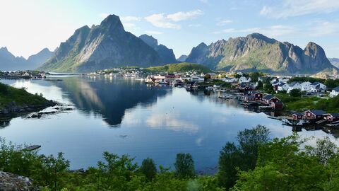 A picturesque coastal village with red and white buildings nestled at the base of dramatic, green-covered mountains, reflected in the calm, blue water.