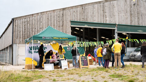 An outdoor event tent for Derbyshire Wildlife Trust, with the slogan "Wilder by nature," is set up in front of a large wooden building, with several people milling around the tent and a car parked in the background.