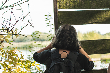 Person with long brown hair, wearing black long sleeve and backpack, watching nature behind a wooden fence 