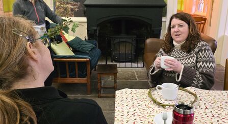 A woman with long brown hair holds a white mug, seated in a brown leather chair, speaking with a woman with blonde hair and glasses in the foreground, while another person places branches near a sofa with green cushions and a fireplace in the background. A table with floral tablecloth has a mug, a spoon, a fork, and a red travel mug.