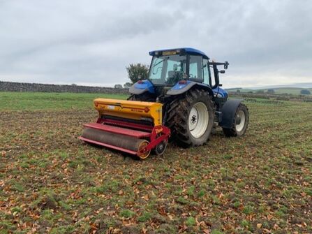 Blue tractor with a yellow and red seeder planting in a field covered in fallen leaves under an overcast sky, with a stone wall and green hills in the background.