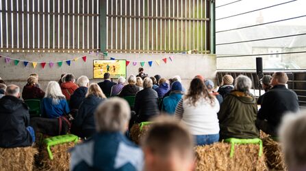 People seated on hay bales listen to a speaker at a meeting inside a barn; a screen displays a logo and image, and colorful pennants hang across the wall.