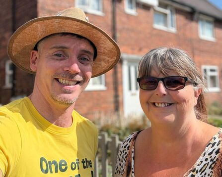 A man wearing a straw hat and a yellow t-shirt, and a woman wearing sunglasses and a leopard-print top smile at the camera in front of a brick house.