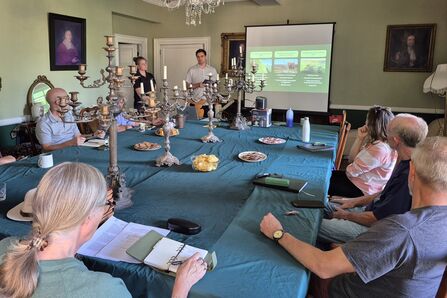 Group of people seated around a large table, attending a presentation projected onto a screen.