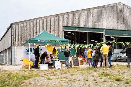 An outdoor event tent for Derbyshire Wildlife Trust, with the slogan "Wilder by nature," is set up in front of a large wooden building, with several people milling around the tent and a car parked in the background.