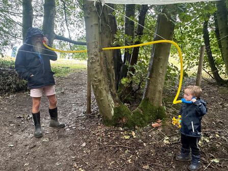 An adult and a young child play with a yellow tube connected between two trees, with the adult holding one end to their ear and the child holding the other.