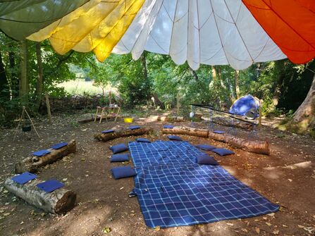 An outdoor forest clearing set up as a nature play area, featuring a colorful parachute canopy overhead, log seating with cushions, a checkered blanket, a small play kitchen, and a blue tent.