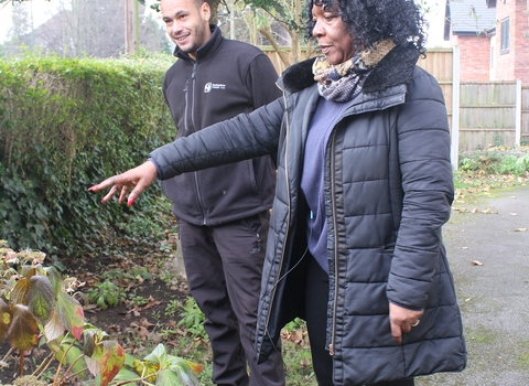a man and a woman stand in a garden, the woman is pointing towards plants near the hedge