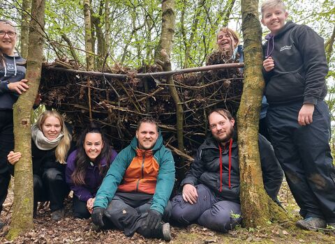 four people sat in a shelter made of sticks with three other stood either side, all are smiling and laughing